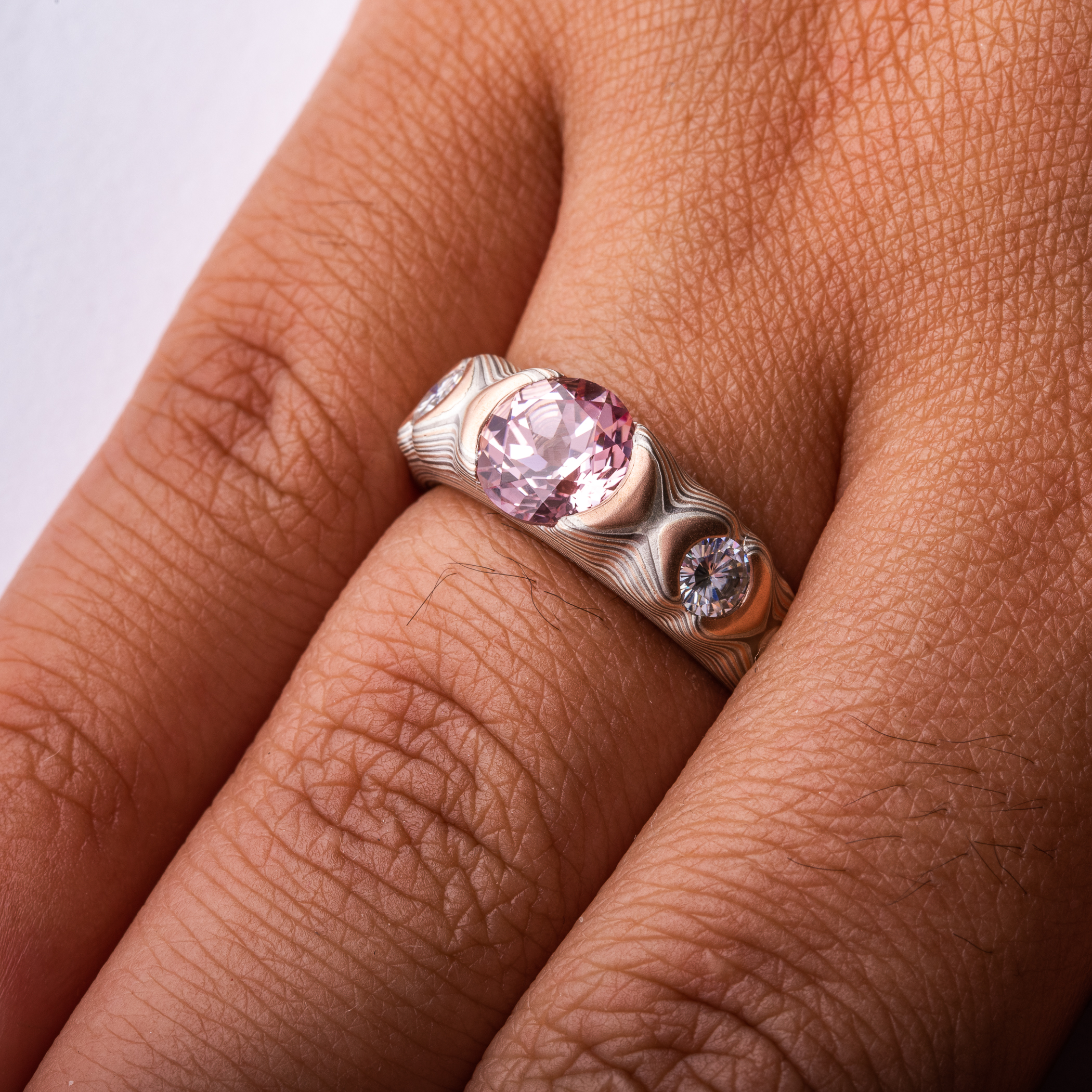 Tapered red gold, palladium and silver mokume gane patterned ring, cathedral style setting with a large pink stone, flanked on each side by a smaller white stone.