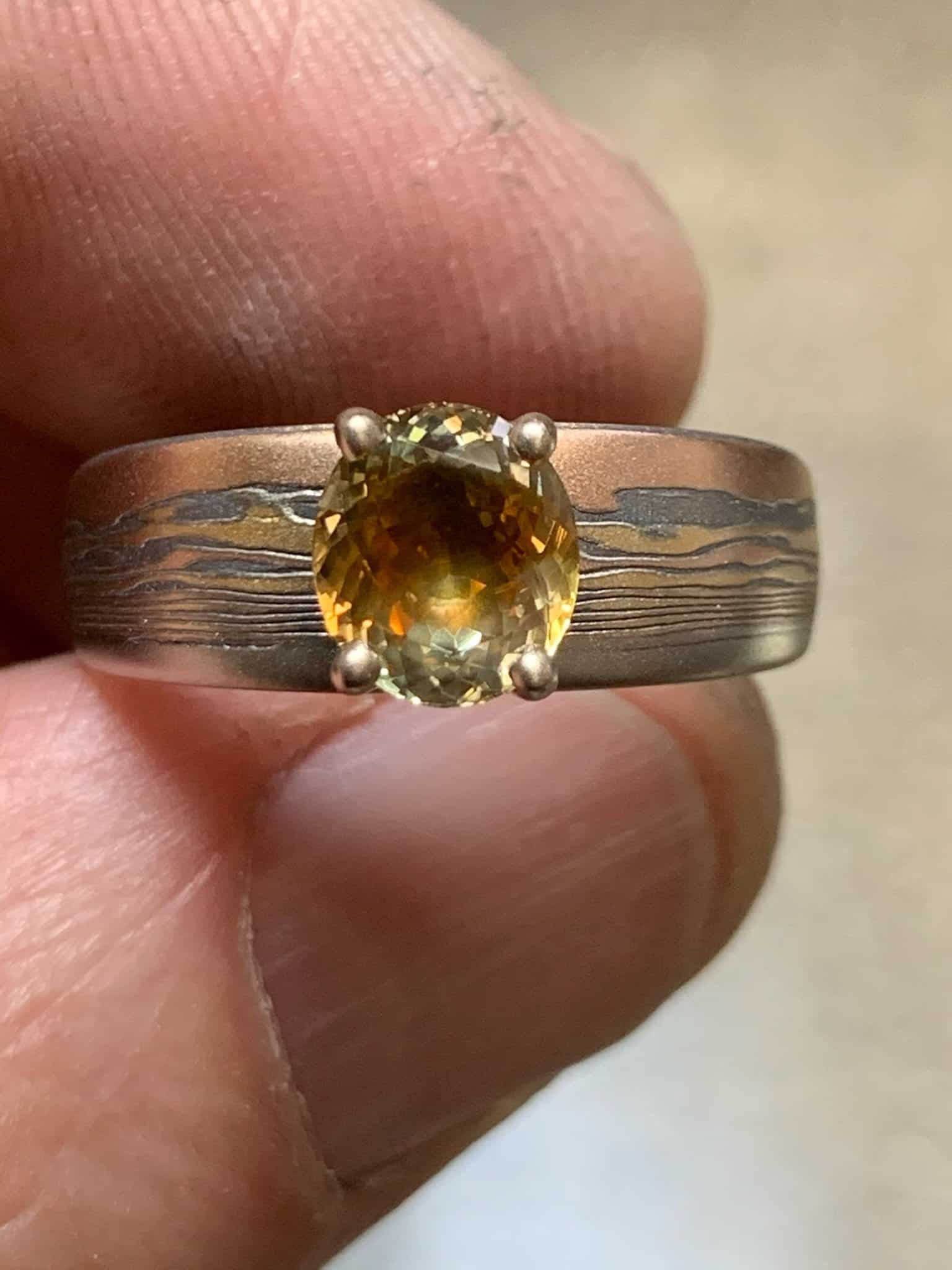 mokume gane ring made with layers of red gold, yellow gold, and oxidized silver, with an orange/yellow oval shaped sapphire set into the band with prongs
