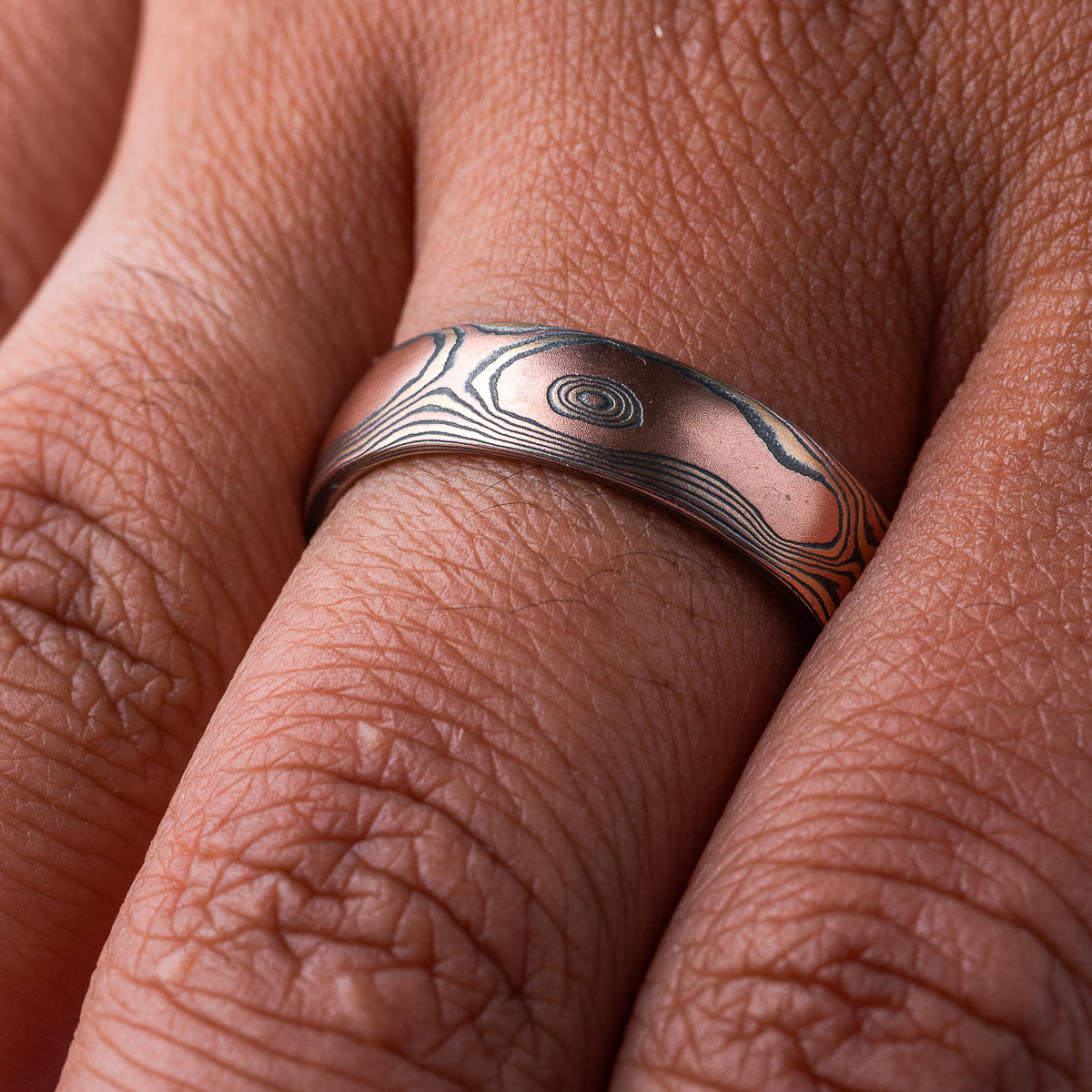 Warm toned mokume gane ring, patterned to look like woodgrain, primarily red gold in color, with yellow gold and oxidized silver (black).