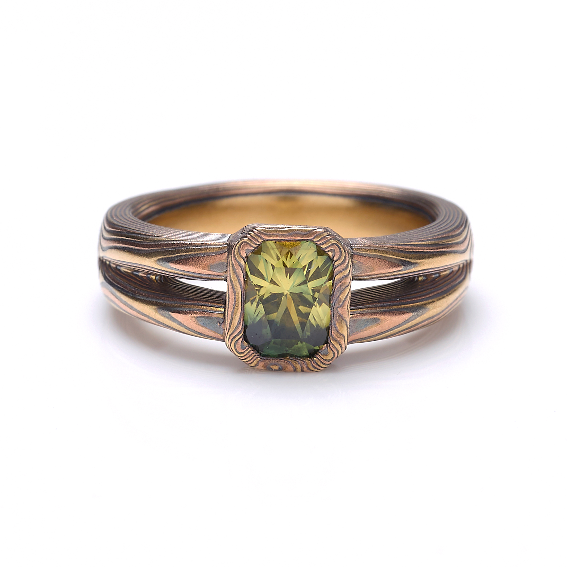 Mokume gane engagement ring with a split shank tapered towards the back of the ring, and a single green emerald cut sapphire placed at the front, with a rim of patterned metal around it.